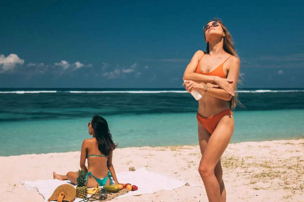 Frau im Bikini trägt am Strand Sonnencreme auf, während eine weitere Frau am Meer sitzt