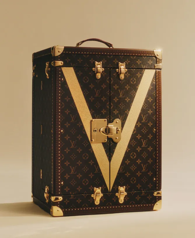 Geschlossener Louis-Vuitton-Koffer mit Monogramm, goldfarbenen Beschlägen und großem cremefarbenem V auf beigem Hintergrund