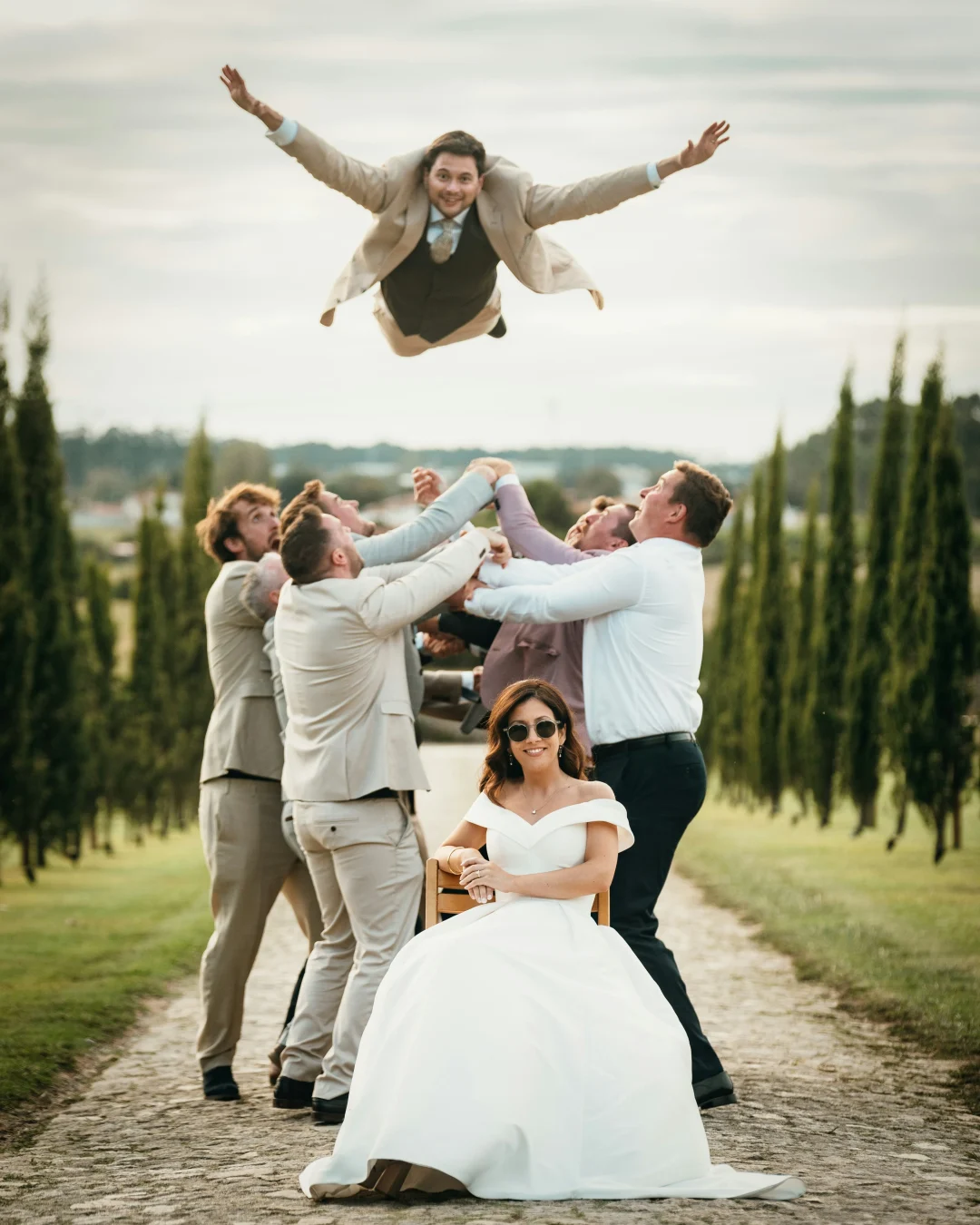 Braut sitzt entspannt im Hochzeitskleid, während Freunde den Bräutigam lachend in die Luft werfen – lebendige Hochzeitsfotografie im Freien
