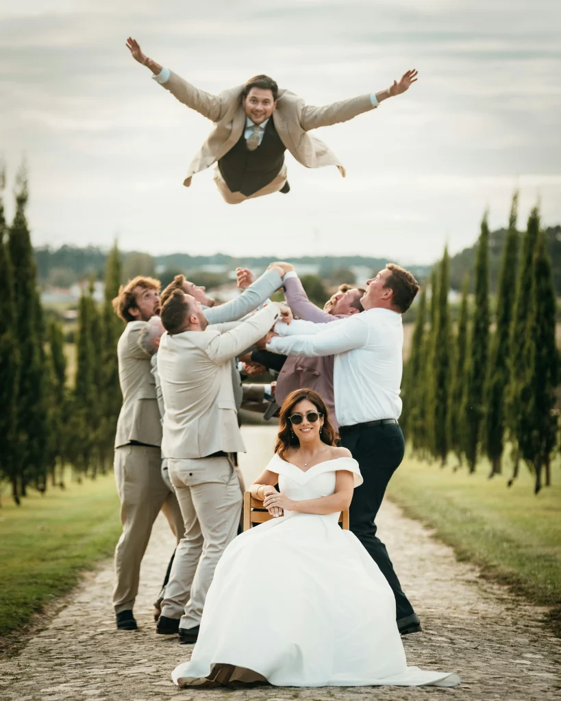 Braut sitzt entspannt im Hochzeitskleid, während Freunde den Bräutigam lachend in die Luft werfen – lebendige Hochzeitsfotografie im Freien