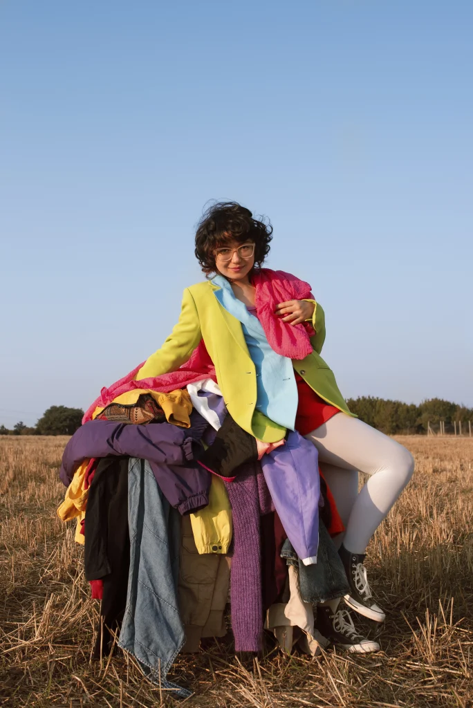 Person sitzt auf einem großen Stapel bunter Kleidung als Symbol für Fast Fashion und Überkonsum