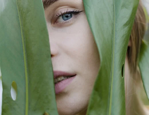 Frau mit strahlender Haut blickt durch grüne Blätter – natürliche Schönheit und frischer Glow