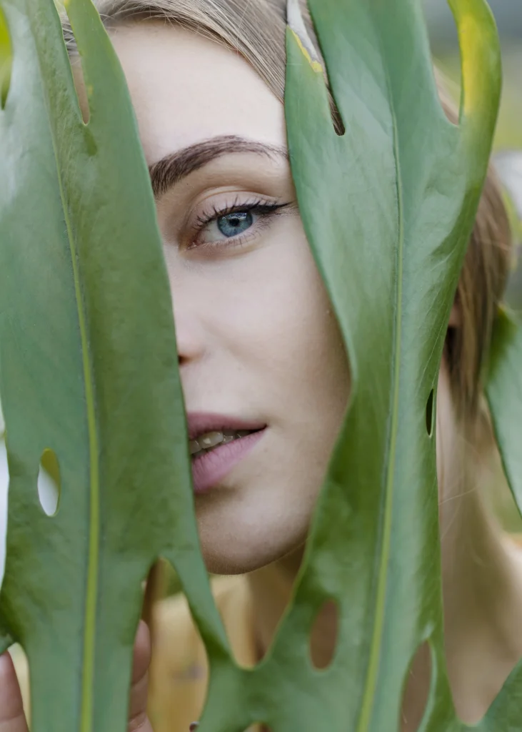 Frau mit strahlender Haut blickt durch grüne Blätter – natürliche Schönheit und frischer Glow