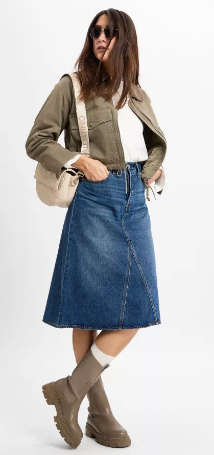 Frau trägt A-Linien-Jeansrock Outfit 2025 – dunkelblauer Denimrock mit khakifarbener Jacke, weißem Oberteil, Tasche und Boots.