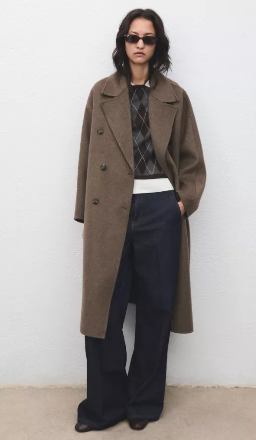 Mantel Damen in Braun – doppelreihiger Wollmantel im Oversize-Schnitt über Argyle-Pullover, kombiniert mit dunkler Wide-Leg-Jeans und braunen Schuhen, vor hellem Hintergrund.