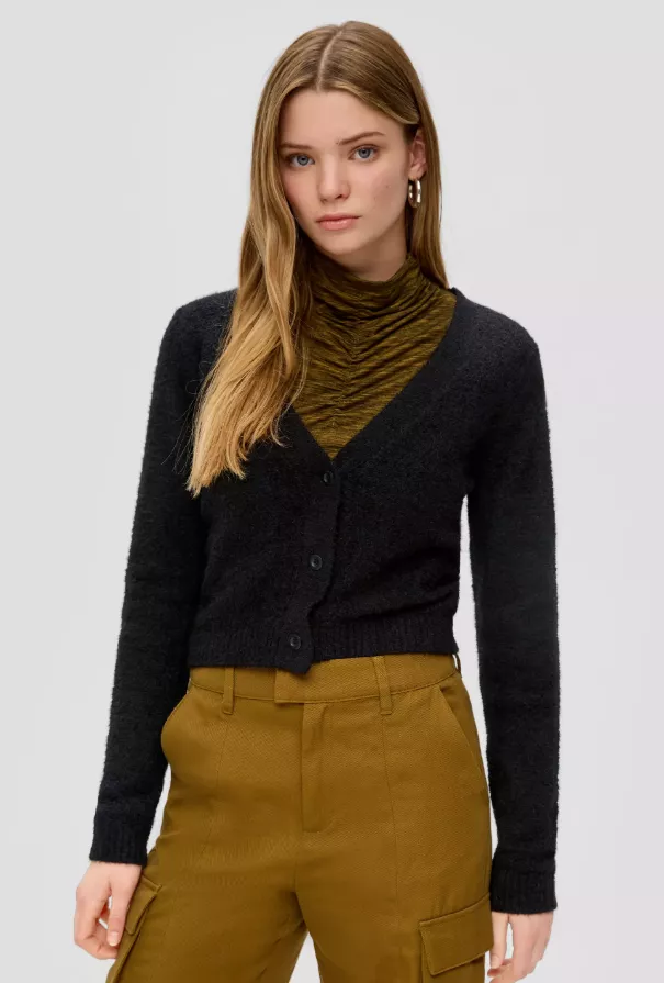 Cardigan vs. Strickjacke – Frau trägt kurzen schwarzen Cropped Damen-Cardigan über olivgrünem Rollkragen und brauner Stoffhose