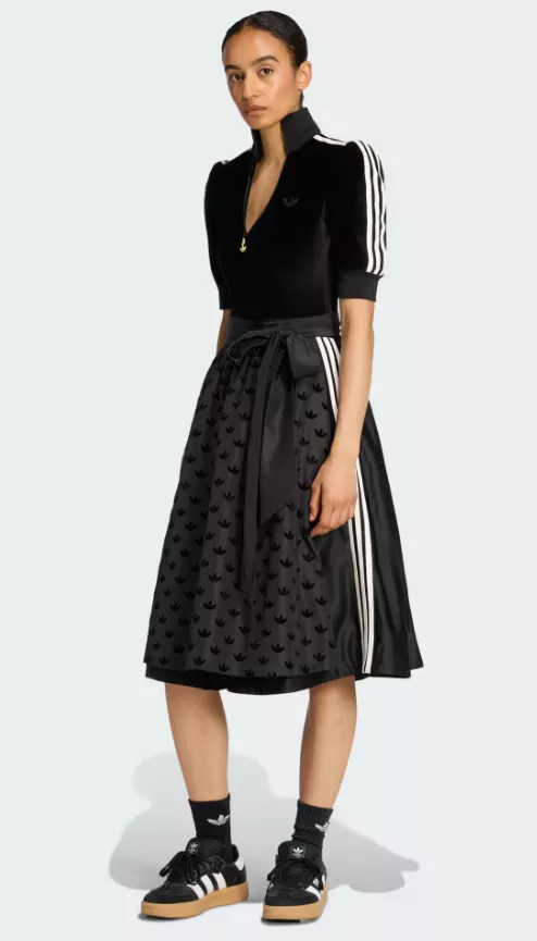 Frau trägt das adidas Dirndl in Schwarz mit dezenten weißen Streifen, Schürze mit Allover-Logo und sportlicher Silhouette.