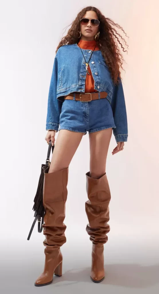 Frau trägt braune Slouchy Overknee Stiefel mit Blockabsatz, Jeansshorts, Jeansjacke und orangem Rollkragenpullover – Herbstmode 2025.