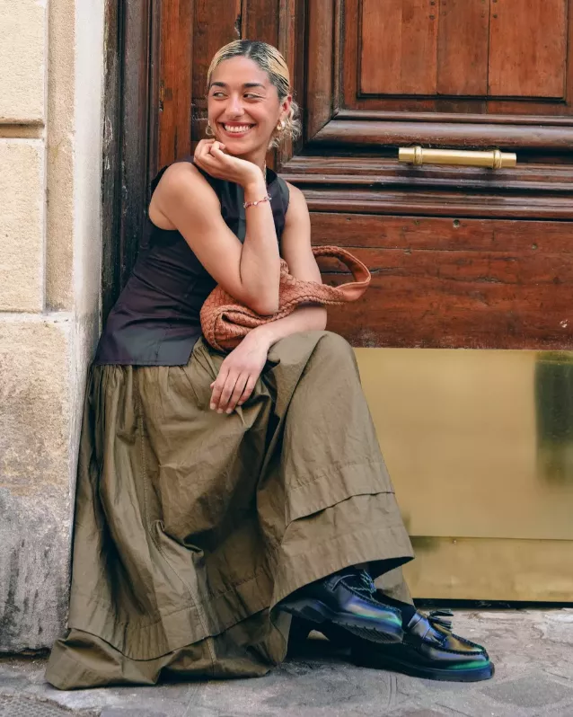 Frau im Streetstyle-Outfit mit langem Rock, ärmellosem Top und schwarzen Loafern mit auffälligem Glanzfinish.