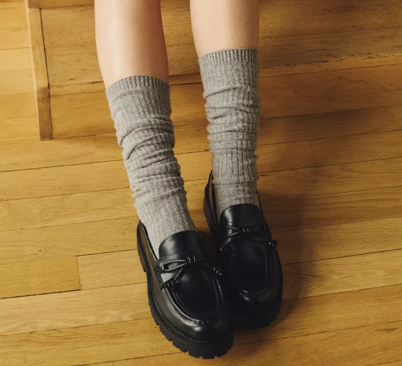 Schwarze Damen-Loafer aus Leder, kombiniert mit grauen, gerippten Socken auf Parkettboden.