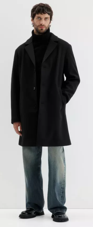 Mantel Herren in Schwarz – einreihiger City-Coat mit Revers über schwarzem Rollkragen, kombiniert mit weiter Jeans und robusten Boots vor hellem Hintergrund.