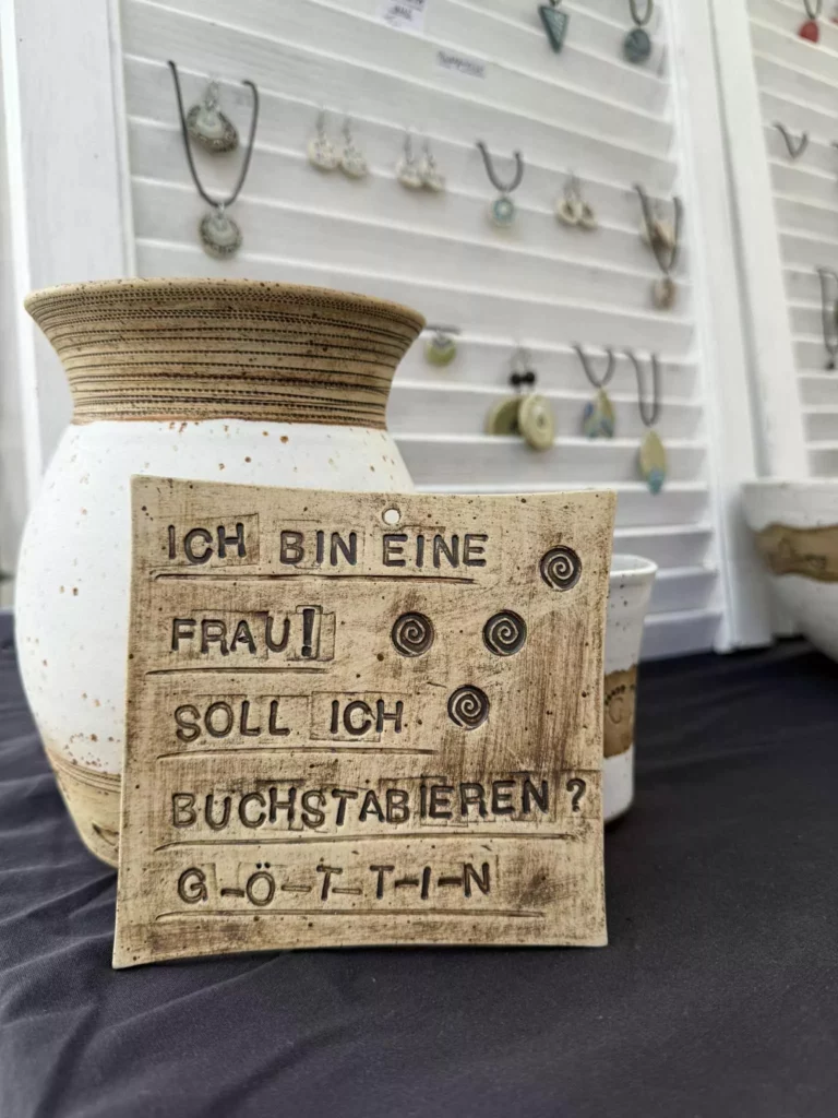 Keramiktafel mit der Aufschrift ‚Ich bin eine Frau! Soll ich buchstabieren? Göttin‘ auf dem Töpfermarkt Neu-Ulm 2025, präsentiert zwischen Vasen und Schmuck.