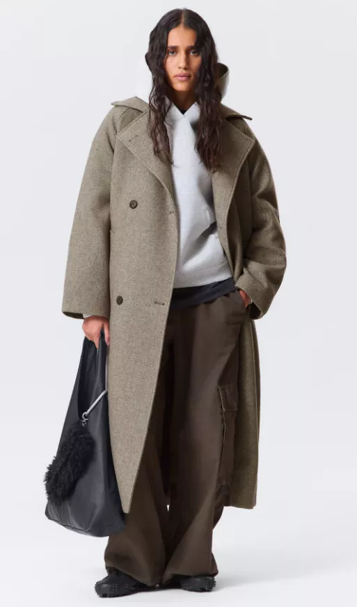 Mantel Damen grau-beige – langer, doppelreihiger Wollmantel im Oversize-Schnitt über grauem Hoodie, kombiniert mit weiten braunen Hosen und großer schwarzer Tote.