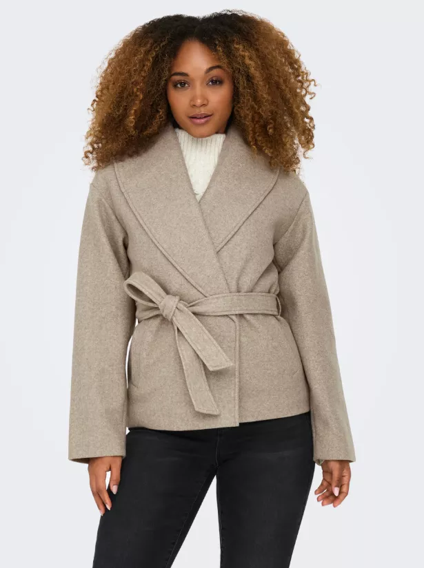 Mantel Damen beige – kurzer Wickelmantel mit Schalkragen und Bindegürtel über cremefarbenem Rippstrickpullover, kombiniert mit schwarzer Jeans vor hellem Hintergrund.