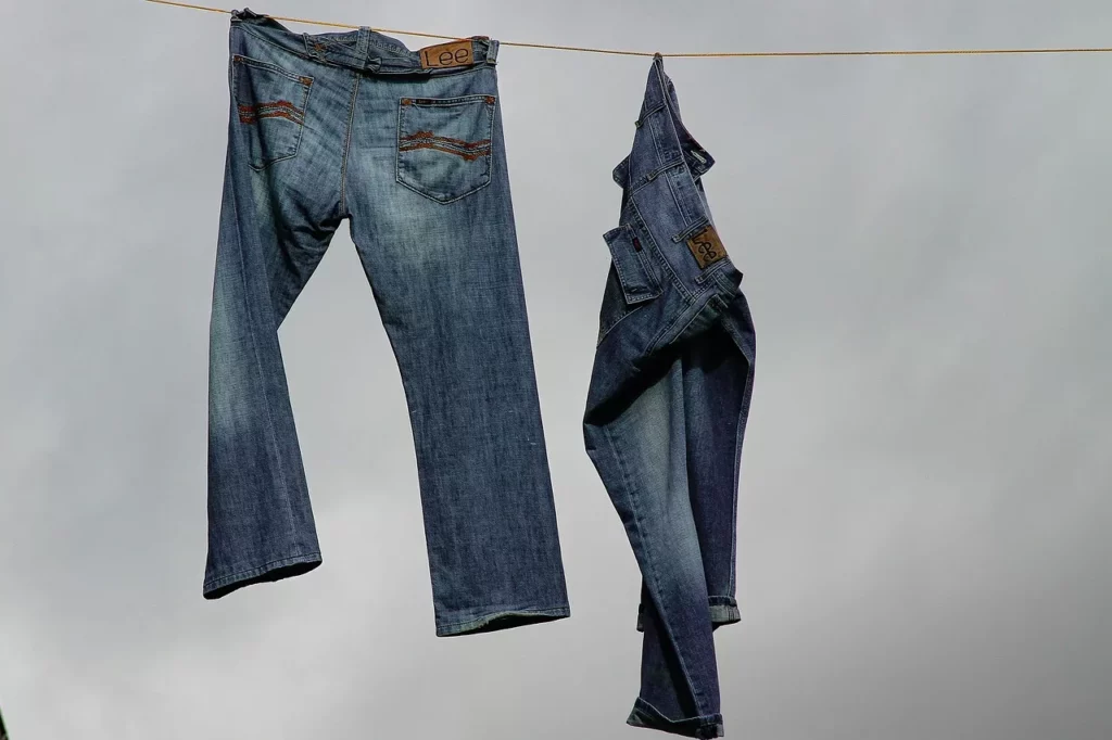 Jeans waschen – zwei blaue Jeans hängen zum Lufttrocknen auf einer Wäscheleine, mit Lederpatch am Bund, wolkiger Himmel im Hintergrund.