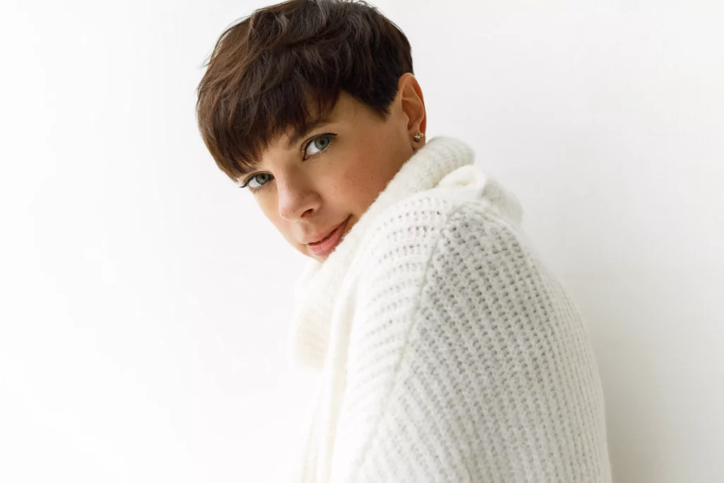 Frau mit kurzem braunen Pixie Cut und weißem Strickpullover vor hellem Hintergrund