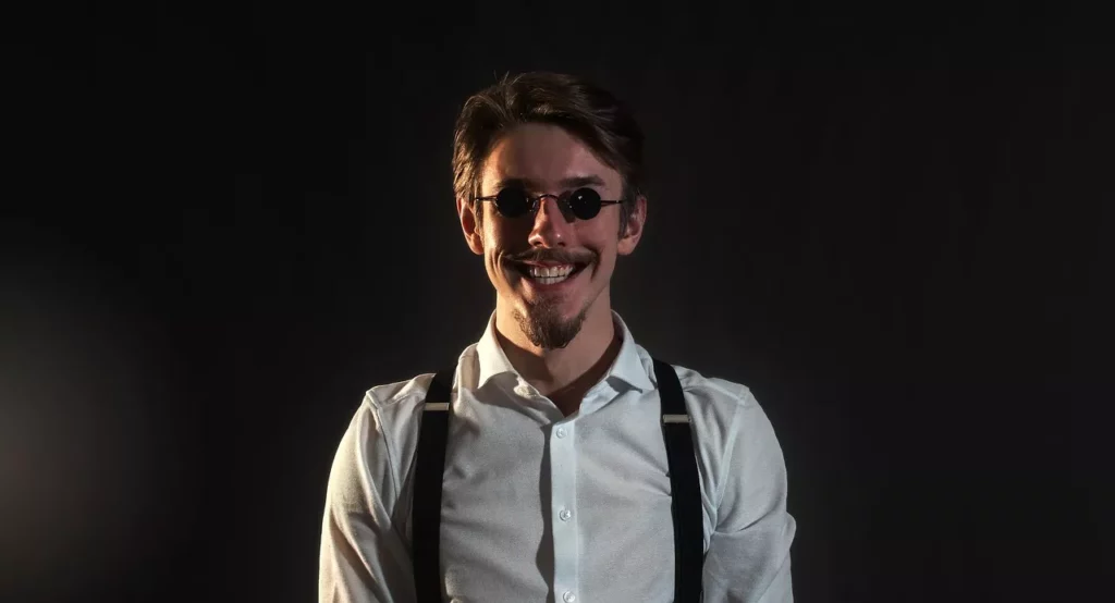 Mann mit Schnurrbart, Spitzbart und Retro-Outfit mit Hosenträgern, lächelnd vor dunklem Hintergrund
