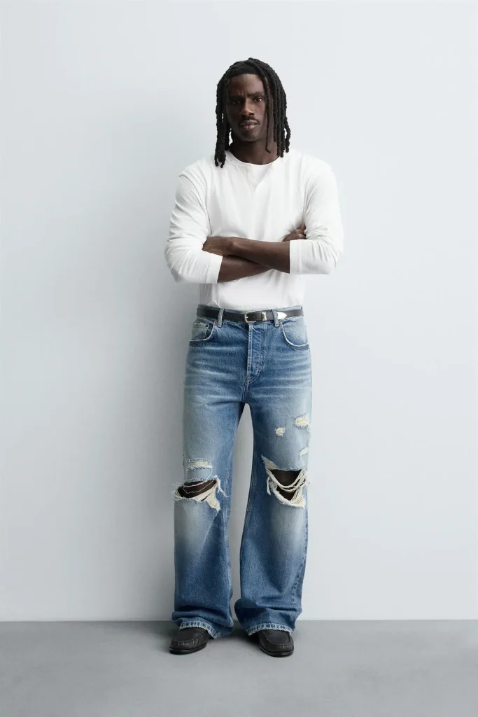 Mann im weißen Longsleeve trägt weit geschnittene ripped jeans in Mittelblau mit großen Knie-Rissen und steht vor neutralem Studiohintergrund.