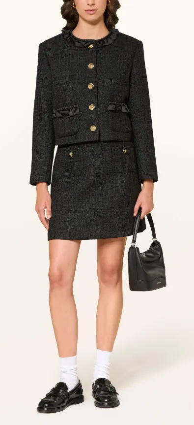 Schwarze Tweed-Jacke mit Volantkragen, goldenen Knöpfen und Schleifendetails, kombiniert mit passendem Minirock, Loafern, Socken und Handtasche.