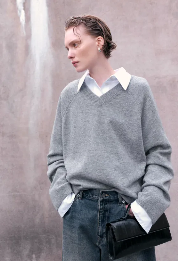 Grauer V-Ausschnitt-Pullover aus Feinstrick über weißem Hemd, kombiniert mit locker sitzender Jeans und schwarzer Clutch – cleaner Herbst-Streetstyle.