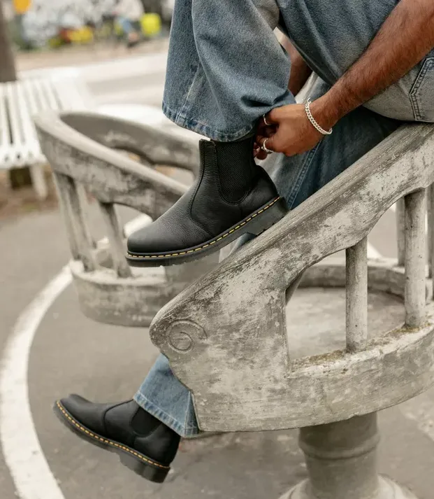 Herrenlook mit schwarzen Chelsea Boots im Dr.-Martens-Stil mit gelber Rahmennaht zu relaxter Jeans auf Steinbank