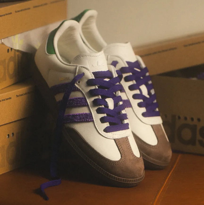 Adidas Samba in Weiß mit braunen Wildleder-Overlays, violetten drei Streifen und passenden lila Schnürsenkeln, grünem Fersenpatch und Gumsohle neben Schuhkartons.