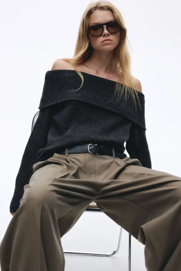 Grauer Off-Shoulder-Pullover aus Rippstrick mit breitem Umschlagkragen, kombiniert mit weit geschnittener taillierter Hose in Olivbraun und schwarzem Ledergürtel; Sitzpose mit eckiger Sonnenbrille.