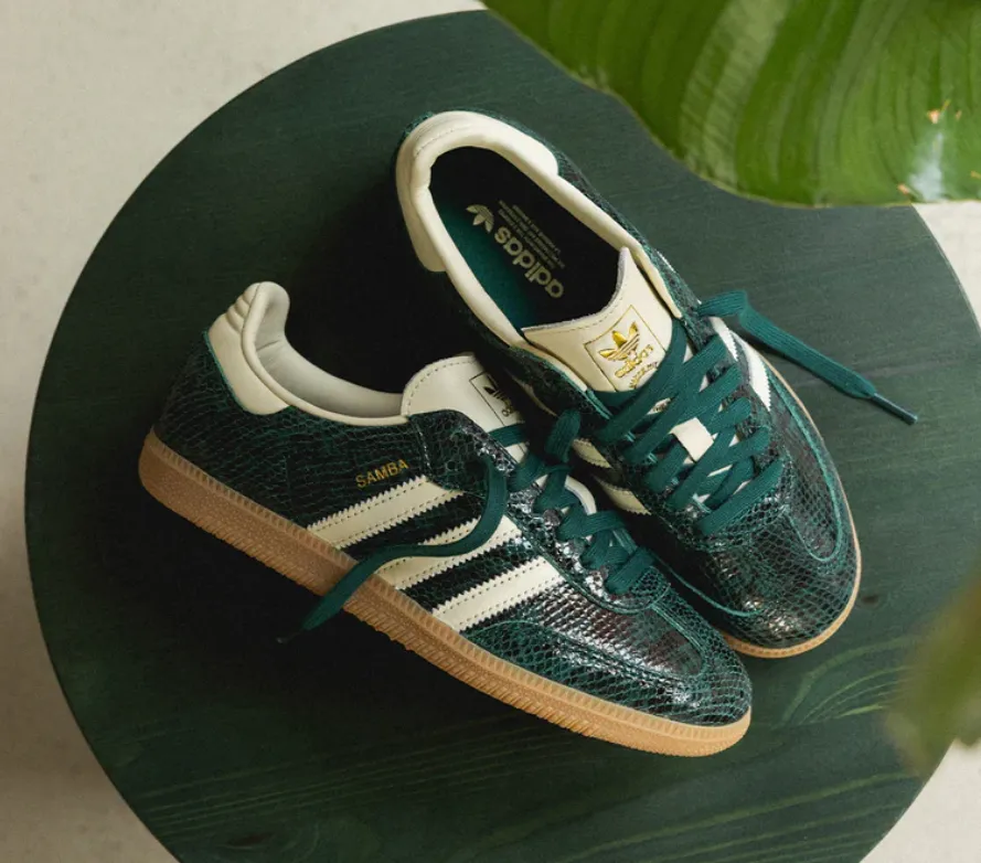 Adidas Samba in Dunkelgrün mit reptilartiger Struktur, weißen drei Streifen, goldnem Zungenlabel und honigfarbener Gumsohle auf rundem, grünem Tisch.