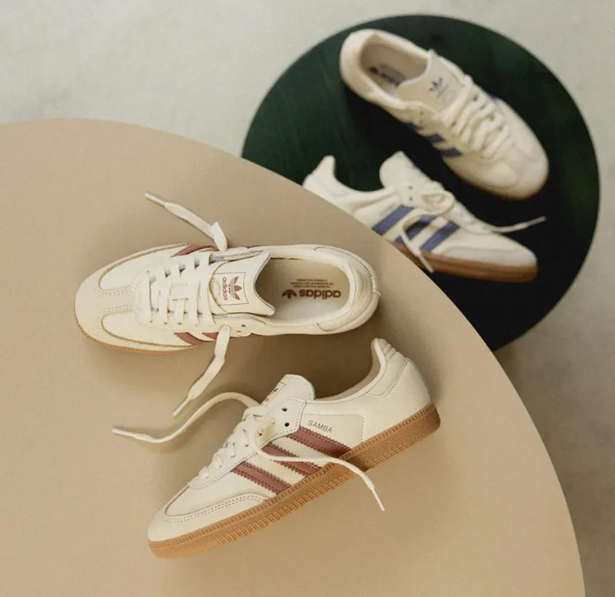 Adidas Samba in Creme mit braunen drei Streifen und Gumsohle, daneben Variante in Creme mit blauen Streifen auf runden Tischen arrangiert.