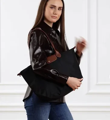 Frau trägt eine schwarze Longchamp Le Pliage Tote mit braunen Lederhenkeln über der Schulter; dunkles Hemd, Jeans, weißer Wandhintergrund.