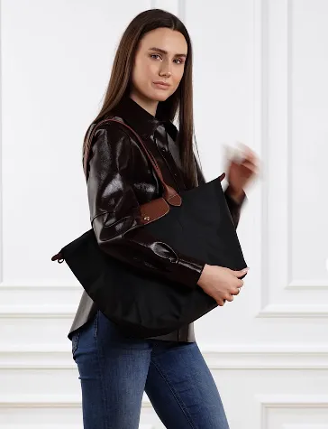 Frau trägt eine schwarze Longchamp Le Pliage Tote mit braunen Lederhenkeln über der Schulter; dunkles Hemd, Jeans, weißer Wandhintergrund.