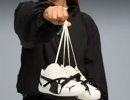 PUMA x ROSÉ Speedcat Lea Sneakers in Creme mit schwarzem Formstreifen und Schleifen-Details, vom Model in schwarzer Jacke vor grauem Hintergrund gehalten.