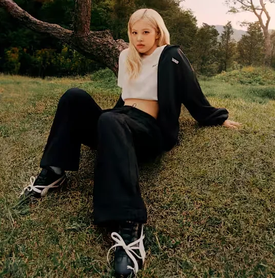 Model in PUMA Outfit sitzt im Gras und trägt schwarze PUMA x ROSÉ Speedcat Sneakers mit weißen Schleifenbändern, Outdoor-Setting bei Sonnenuntergang.