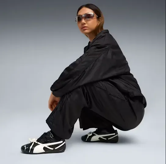 Model in schwarzem Oversize-Set hockt vor grauem Hintergrund und trägt schwarze PUMA x ROSÉ Speedcat Sneakers mit kontrastierendem weißen Formstreifen und Schleifen.