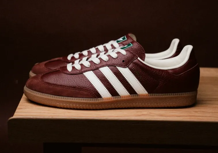 Adidas Samba in dunkelbraun mit weißen drei Streifen und Gummisohle auf einem Holztisch, seitliche Ansicht, grünes Zungenlabel.