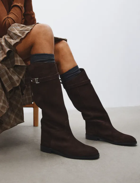 Dunkelbraune, flache Wildlederstiefel in Kniehöhe mit schmaler Schnalle, kombiniert mit kariertem Midirock und gerippten Socken.