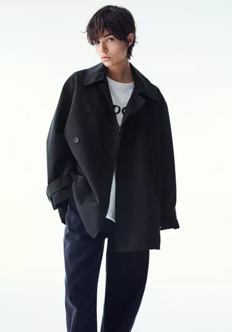 Damen Trenchcoat kurz in Schwarz, oversized, doppellagige Knopfleiste, über weißem T-Shirt und dunkler, weiter Hose im Studio.