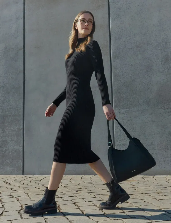 Frau im schwarzen Rippstrick-Midikleid trägt schwarze Chelsea Boots mit Profilsohle und geht vor Betonwand – monochromer Herbstlook