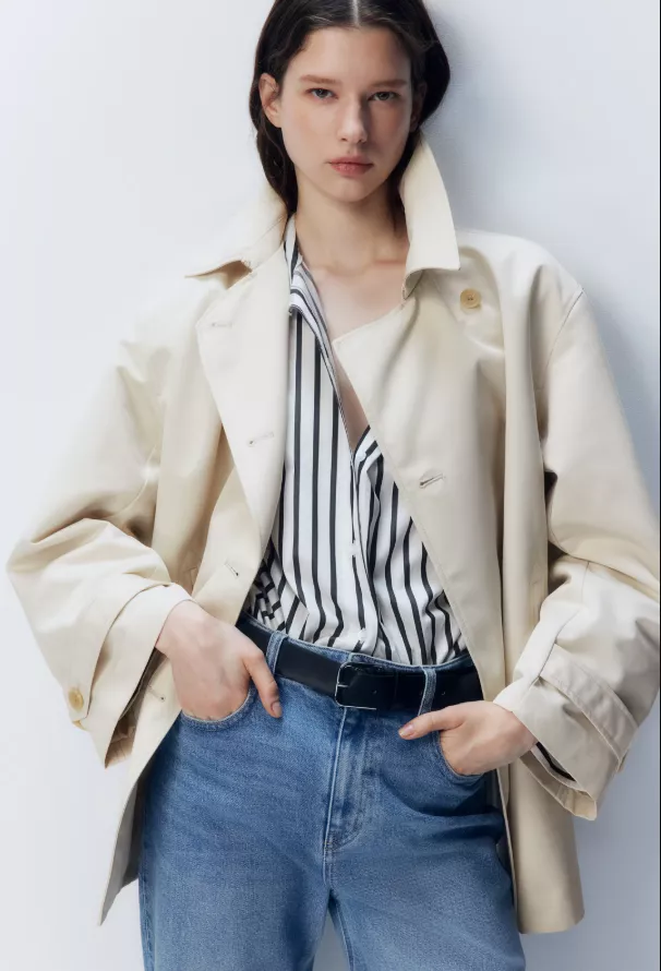 Damen Trenchcoat kurz in Beige, locker geschnitten, über gestreifter Bluse und Mid-Rise-Jeans, Hände in den Taschen.