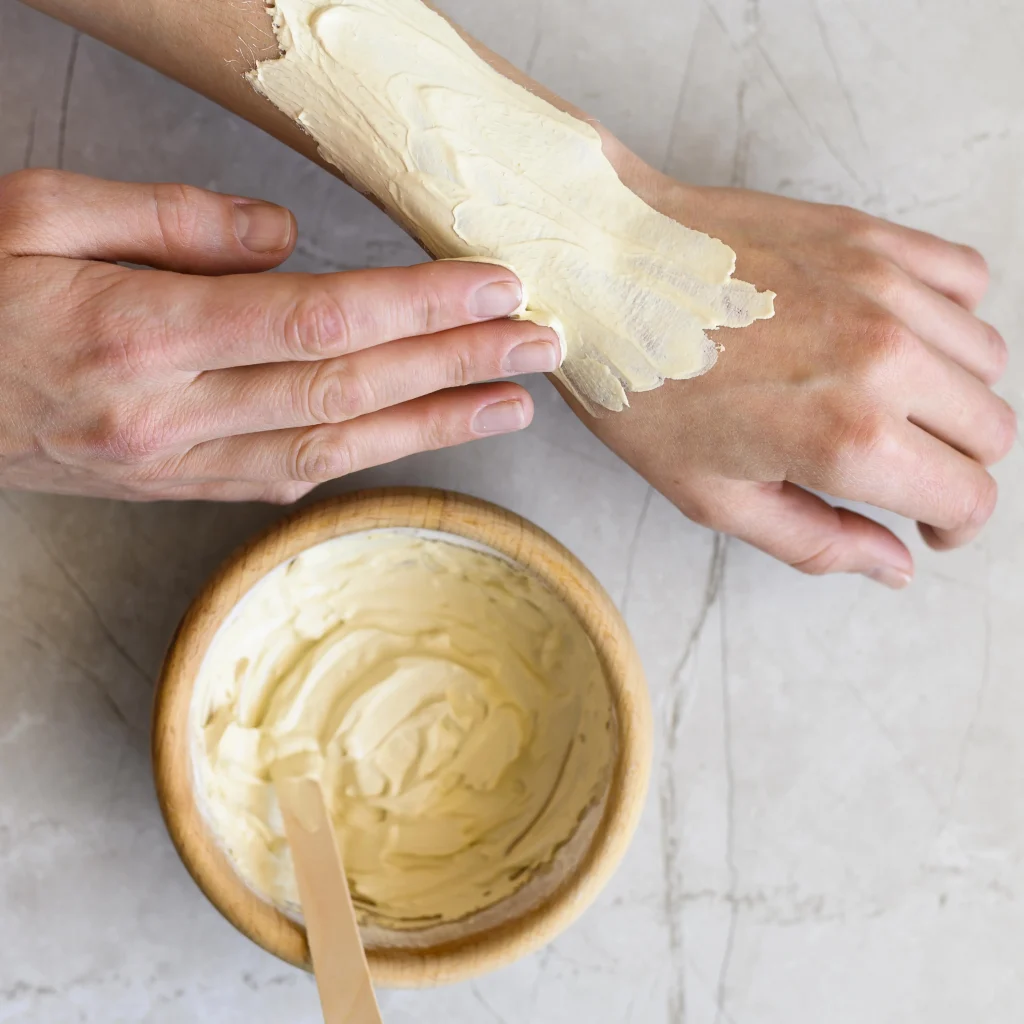 Hände tragen cremige tallow-Creme auf den Unterarm auf, Holzschale mit Balm und Spatel auf hellem Steinuntergrund