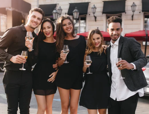 Gruppe junger Erwachsener in eleganter Dresscode-Cocktail-Kleidung, schwarze Midikleider und dunkler Blazer, mit Weingläsern auf einer Stadtterrasse am frühen Abend.