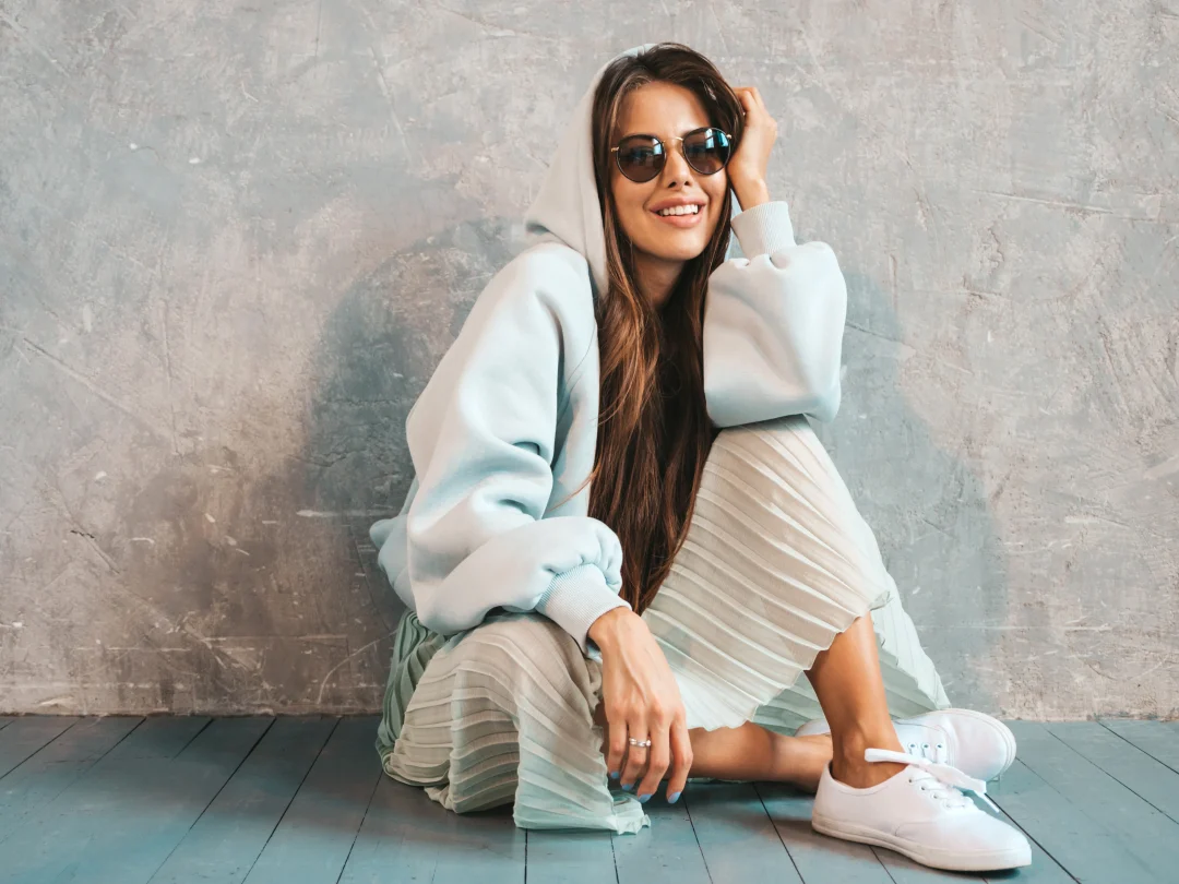 Casual chic Look: Frau mit pastellfarbenem Hoodie, plissiertem Midirock und weißen Sneakern, sitzend vor grauer Betonwand, mit Sonnenbrille
