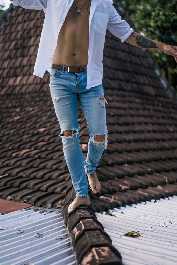 Mann mit offener weißer Hemdjacke balanciert barfuß auf einem Ziegeldach und trägt hellblaue ripped jeans mit Knie-Cut-outs.