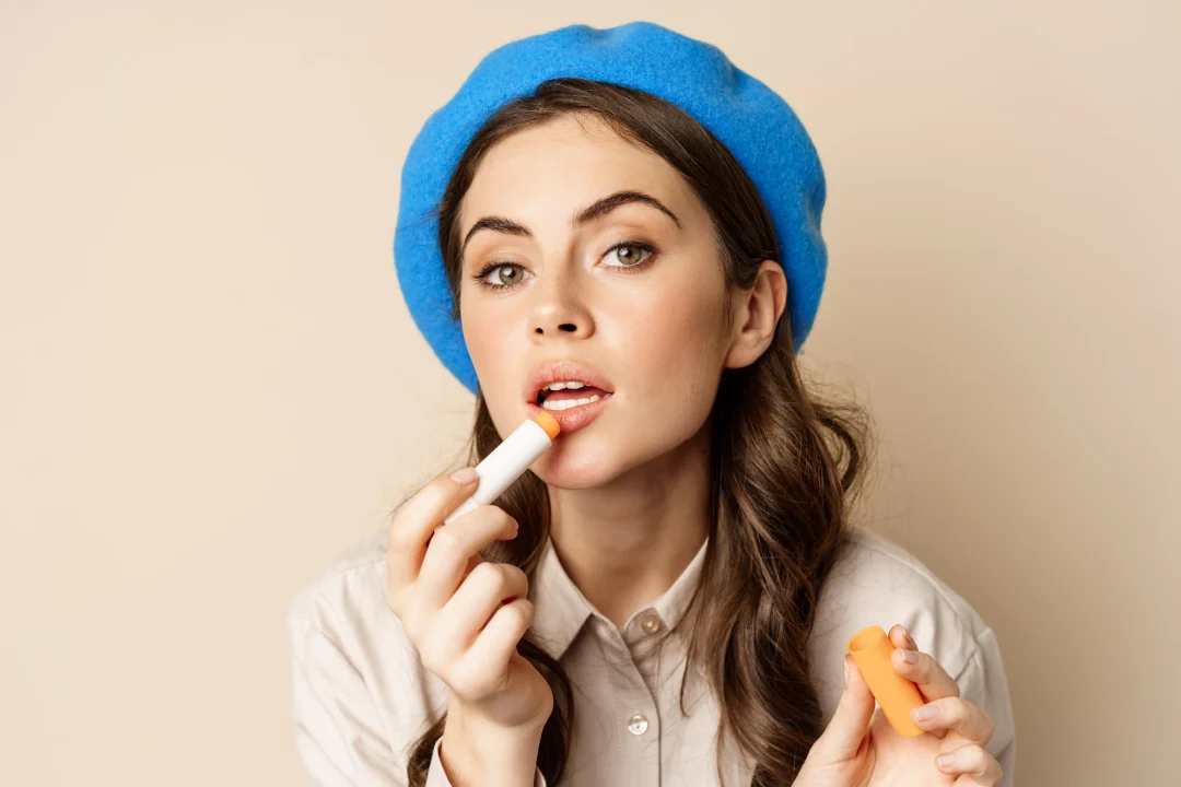 Lippenpflege im Alltag – junge Frau trägt Lippenbalsam auf, Nahaufnahme vor beigem Hintergrund, dezentes Make-up, blauer Barett, natürlicher Glow