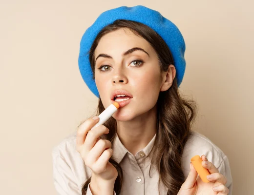 Lippenpflege im Alltag – junge Frau trägt Lippenbalsam auf, Nahaufnahme vor beigem Hintergrund, dezentes Make-up, blauer Barett, natürlicher Glow