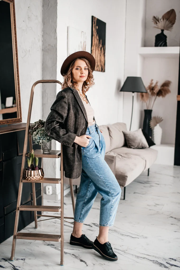 Frau im Casual-chic Look mit Blazer, Jeans, Hut und schwarzen Loafern in modernem Wohnzimmer-Setting