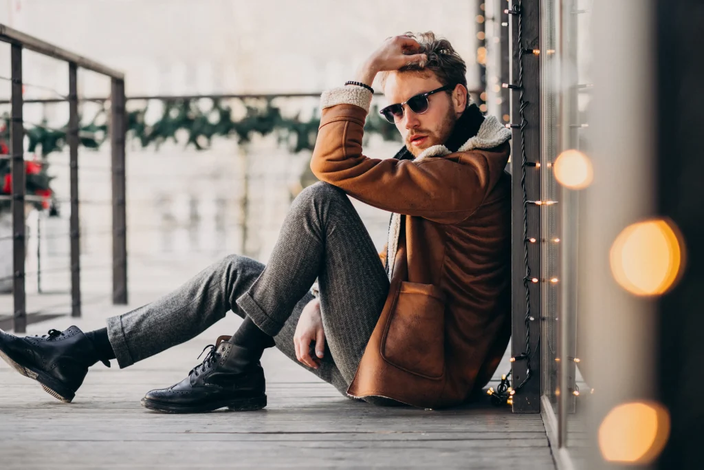 Mann im Casual-chic Look mit brauner Shearling-Jacke, grauer Wollhose und schwarzen Boots, sitzend auf Holzsteg mit Lichterkette