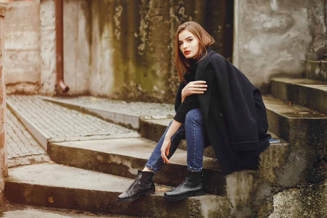 Frau im dunkelblauen Mantel und Skinny-Jeans trägt schwarze Chelsea Boots und sitzt auf Treppen im urbanen Herbst-Setting