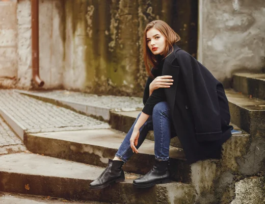 Frau im dunkelblauen Mantel und Skinny-Jeans trägt schwarze Chelsea Boots und sitzt auf Treppen im urbanen Herbst-Setting