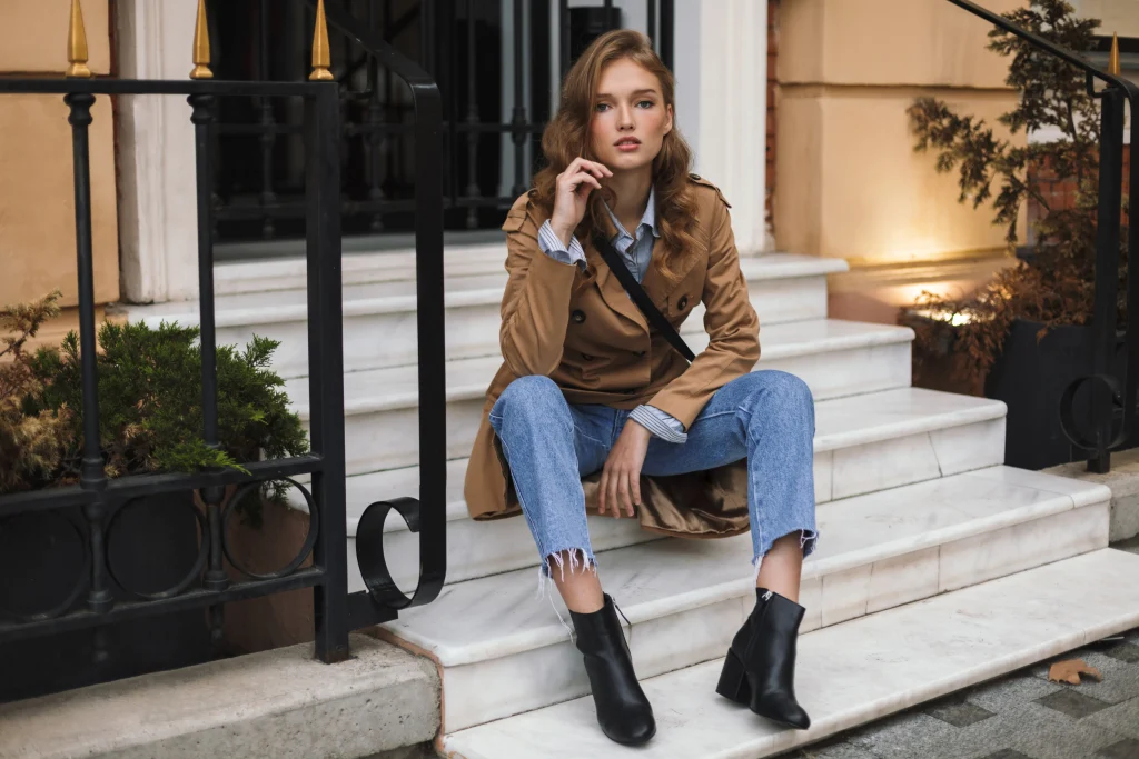 Frau im Trenchcoat sitzt auf Marmorstufen und trägt schwarze Ankle Boots mit Blockabsatz zu hellblauer Jeans